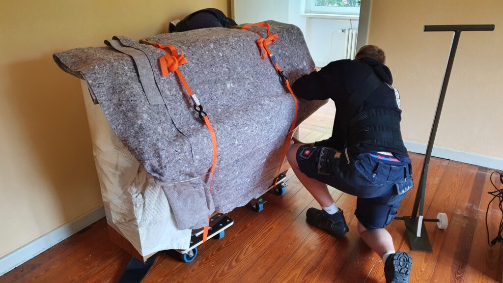 Mitarbeiter von Klaviertransport Lüneburg schützt und sichert ein Klavier auf Rollbrettern